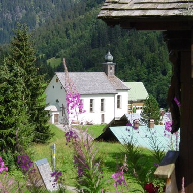 Eine malerische Landschaft mit einer Kirche und bunten Blumen im Vordergrund. Hohe Bäume und sanfte Hügel umgeben die Szene. | © Kleinwalsertal Tourismus | Rolf Köberle