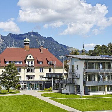 Ein charmantes Hotelgebäude mit rotem Dach und moderner Architektur. Im Hintergrund sind Berge und ein blauer Himmel zu sehen. | © Klinikverbund Allgäu