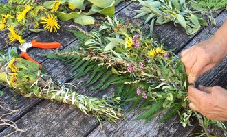 Ein Tisch mit frischen Kräutern und bunten Blumen. Jemand bindet einen Kräuterkranz mit einer Schere daneben. | © Kleinwalsertal Tourismus | Lydia Fritz-Ilg