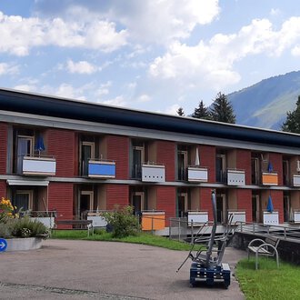 A modern building with several floors and balconies. In the background, green hills and a blue sky can be seen. | © Kleinwalsertal Tourismus | Veronika Senn