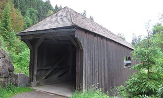 Eine alte Holzbrücke inmitten eines grünen Waldgebiets. Die Brücke hat ein spitzes Dach und ist von Bäumen umgeben. | © Stefan Heim