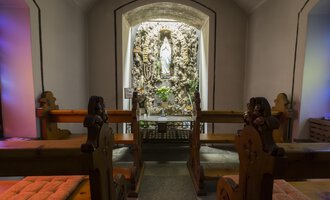 Eine stille Kapelle mit Holzbänken und einer Statue in der Nische. Die Wände sind sanft beleuchtet und schaffen eine friedliche Atmosphäre. | © Kleinwalsertal Tourismus | Oliver Farys