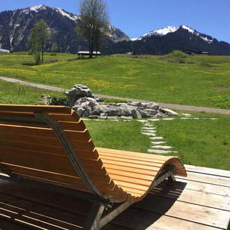 A wooden lounge chair is on a terrace overlooking green meadows and snow-capped mountains in the background. The sky is clear and the landscape looks idyllic. | © Kleinwalsertal Tourismus | Sarina Berchtold