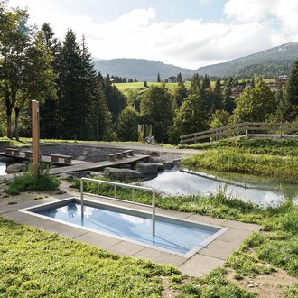 Ein kleiner Pool in einer grünen Umgebung mit Bäumen und einer ruhigen Wasserfläche im Hintergrund. Die Landschaft ist sanft hügelig und lädt zur Entspannung ein. | © Kleinwalsertal Tourismus | Veronika Senn