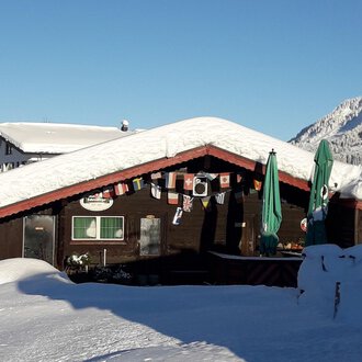 A cozy wooden house with a snow-covered roof in a wintry landscape. In the background, there are snow-covered mountains and a blue sky. | © Panoramahütte