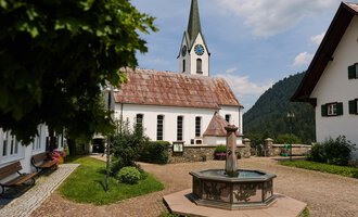 A picturesque village square with a fountain in the foreground and traditional houses. In the background, there is a church with a tall tower and a cloudy sky. | © Kleinwalsertal Tourismus | Oliver Farys