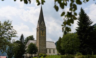 A picturesque street with a church and a traditional building. In the background, one can see green hills and a blue sky with white clouds. | © Kleinwalsertal Tourismus | Oliver Farys