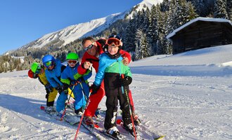 Eine Gruppe von Kindern und einem Erwachsenen beim Skifahren in der verschneiten Berglandschaft. Sie tragen bunte Skikleidung und lächeln in die Kamera. | © Andy Herr | Werbewind GmbH