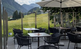 An inviting terrace with black chairs and tables. In the background, green meadows and mountains can be seen under a blue sky. | © Robert Haller GmbH c/o Hotel Steinbock