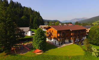 Ein gemütliches Holzhaus liegt in einer grünen Landschaft, umgeben von Bergen und Bäumen. Im Vordergrund befindet sich ein Spielplatz für Kinder. | © Hotel Bellevue | Werbewind GmbH