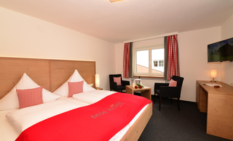 A cozy hotel room with a double bed, two chairs, and a table. The walls are bright, and the room has large windows with curtains.