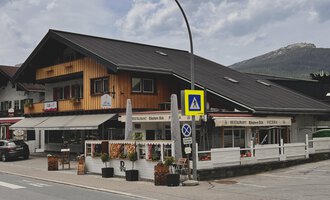 A traditional alpine building with wooden cladding and a restaurant on the ground floor. In the foreground, flower arrangements and traffic signs can be seen. | © Riezlern Eck | Sead Sulejmani