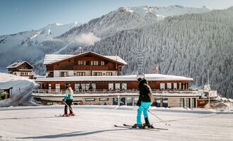 Zwei Skifahrer fahren auf einer verschneiten Piste vor einem schönen Berghotel. Die Landschaft ist von Bergen und skandinavischen Wäldern umgeben.