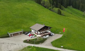 A cozy building in nature with green meadows. Several cars are parked in front of the house, which is surrounded by trees.