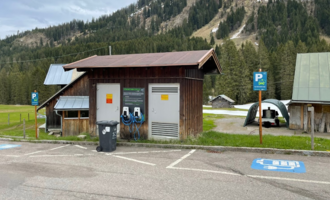 Eine kleine Holzgebäude mit Ladestationen für Elektroautos steht auf einem Parkplatz im Freien. Im Hintergrund sind grüne Wiesen und bewaldete Hügel zu sehen. | © Kleinwalsertal Tourismus