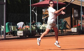 A man is playing tennis on a sand court. In the background, there are chairs and tables as well as a few plants. | © Sportverein Kleinwalsertal