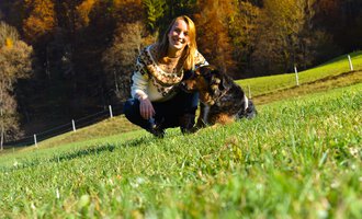 A woman is sitting in the grass and smiling next to a dog. In the background, trees and a green meadow can be seen. | © Tierarztpraxis Oberstdorf | Regina Baumgartner