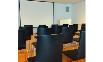 A room with black chairs and a projection screen. The floor is made of wood and the walls are bright.