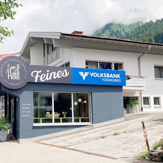 Ein modernes Gebäude mit einem Geschäftsschild „Feines“ und der Volksbank Vorarlberg. Im Hintergrund sind grüne Hügel und ein klarer Himmel zu sehen. | © Volksbank Vorarlberg e. Gen.