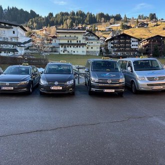 Vier Taxis stehen in einer Reihe vor einer malerischen Berglandschaft. Im Hintergrund sind traditionelle alpine Gebäude und bewaldete Hügel zu sehen. | © Walser Taxi | Josef Düringer