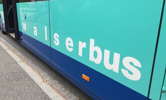 A bus with the inscription "walserbus" in light blue and blue color. The bus is at a stop on a street. | © Kleinwalsertal Tourismus
