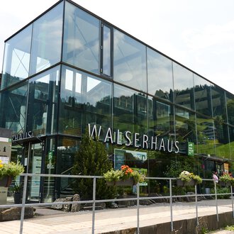 A modern building with large glass windows, surrounded by green plants. It bears the inscription "WALSERHAUS". | © Kleinwalsertal Tourismus | Oliver Farys