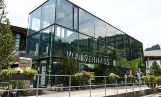 A modern building with large glass windows, surrounded by green plants. It bears the inscription "WALSERHAUS". | © Kleinwalsertal Tourismus | Oliver Farys