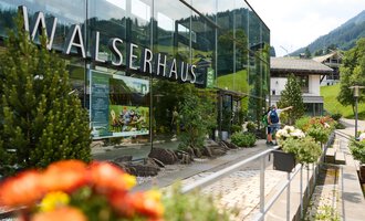 The Walserhaus is a modern building with large windows in a picturesque setting. In the foreground, colorful flowers and a hiker can be seen. | © Kleinwalsertal Tourismus | Oliver Farys