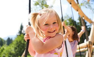 A cheerful girl is swinging on a swing. In the background, trees and another playmate can be seen. | © Kleinwalsertal Tourismus | Oliver Farys