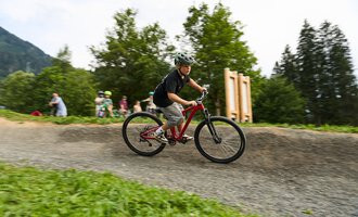 A boy is riding a mountain bike on a winding path. In the background, more children and trees can be seen. | © Kleinwalsertal Tourismus | Oliver Farys