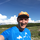 A smiling man in a blue t-shirt and orange cap outdoors in a green landscape with a blue sky and clouds. | © Kleinwalsertal Tourismus eGen