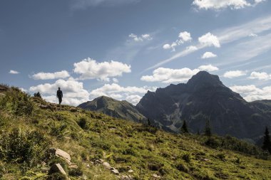 Ein einzelner Wanderer steht auf einer grünen Berghalbinsel mit Blick auf die Berge und den blauen Himmel im Schwarzwassertal. | © Kleinwalsertal Tourismus eGen • Photographer: Andre Tappe