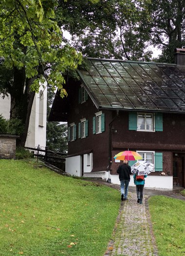 Two people with umbrellas walking on a path past a house in a green landscape. | © Kleinwalsertal Tourismus eGen • Photographer: Andre Tappe
