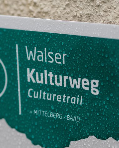Sign for the Walser Culture Trail in Mittelberg-Baad, wet from rain, with water droplets on the surface. | © Kleinwalsertal Tourismus eGen • Photographer: Andre Tappe