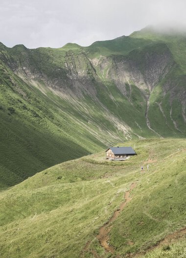 A solitary cabin nestled among green rolling hills with steep, forested mountain slopes in the background. | © Kleinwalsertal Tourismus eGen • Photographer: Andre Tappe