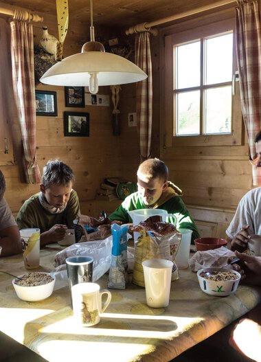 Family and children having breakfast at a wooden table in a cozy, wood-paneled kitchen, illuminated by sunlight. | © Kleinwalsertal Tourismus eGen • Photographer: Andre Tappe