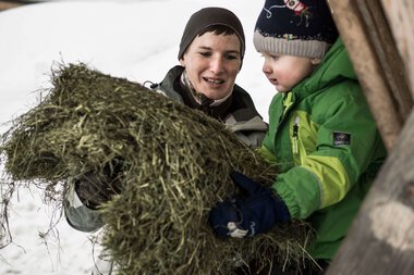 A woman and a child in the snow at a forest feeding scene, holding a big bundle of hay or wild food together. | © Kleinwalsertal Tourismus eGen • Photographer: Andre Tappe
