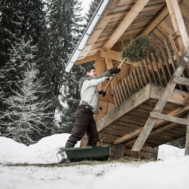 Young person throws hay or branches from a balcony of a wooden house in a snowy forest setting. | © Kleinwalsertal Tourismus eGen • Photographer: Andre Tappe