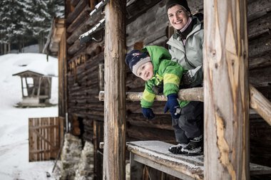 A woman and a child look out of a window of a wooden house in the snow, smiling and enjoying the winter nature. | © Kleinwalsertal Tourismus eGen • Photographer: Andre Tappe