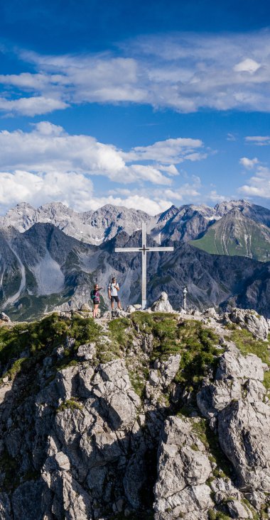 Berggipfel mit Gipfelkreuz und Wanderern, oberhalb der Alpen, bei schönem Wetter, mit blauer Himmel und Wolken. | © Kleinwalsertal Tourismus eGen • Fotograf: Bastian Morell