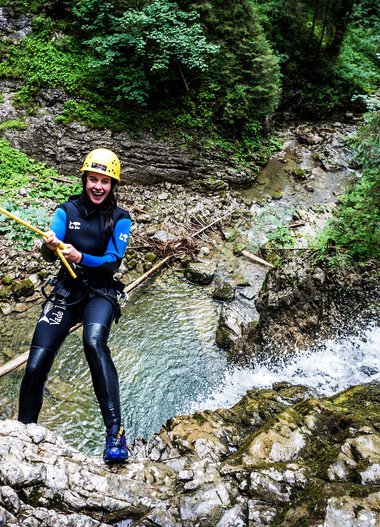 Woman canyoning in a river surrounded by lush green forest, wearing a wetsuit, helmet, and secured with a rope. | ©   Kleinwalsertal Tourismus eGen • Photographer: Dominik Berchtold