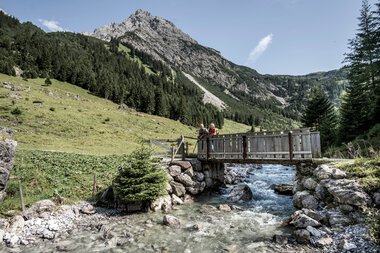 Two people on a wooden bridge over a creek in an alpine landscape with mountains in the background. | © Kleinwalsertal Tourismus eGen • Photographer: Dominik Berchtold