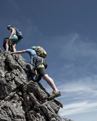 Two climbers ascending a rocky mountain peak, with a third climber helping, under clear skies on a sunny day. | © Kleinwalsertal Tourismus eGen • Photographer: Dominik Berchtold