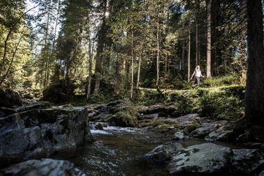 Eine Person wandert entlang eines kleinen Flusses im dichten Wald mit hohen Bäumen, Sonnenlicht filtert durch das Laub. | © Kleinwalsertal Tourismus eGen • Fotograf: Dominik Berchtold