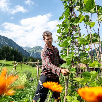 Mann in einem Permakulturgarten, umgeben von blühenden Pflanzen und grünen Bergen im Hintergrund, bei sonnigem Wetter. | © Kleinwalsertal Tourismus eGen • Fotograf: Dominik Berchtold