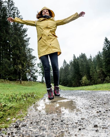 Woman in a yellow raincoat and dark pants standing on a wet forest path with arms spread out in bad weather in the forest. | © Kleinwalsertal Tourismus eGen • Photographer: Dominik Berchtold