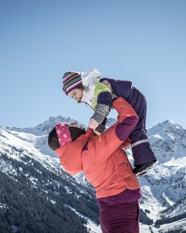 Family winter hiking in the mountains, an adult lifts a child onto a snow mound, with snow-covered peaks under a blue sky in the background. | © Kleinwalsertal Tourismus eGen • Photographer: Dominik Berchtold