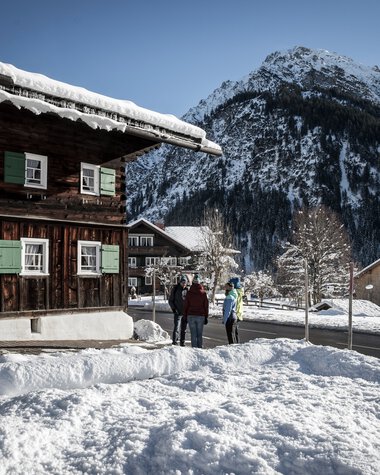 Three people walking on a snow-covered street in a winter village, with mountains in the background. | © Kleinwalsertal Tourismus eGen • Photographer: Dominik Berchtold