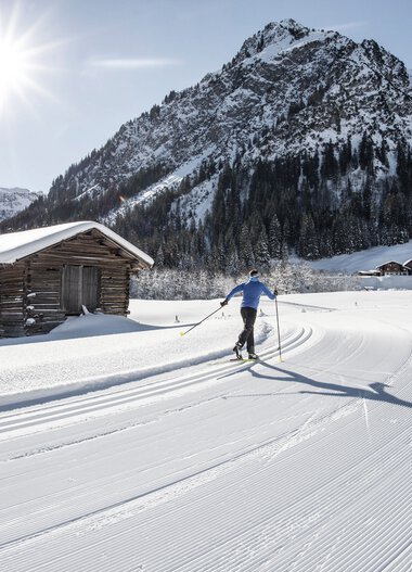 Cross-country skier on a snow-covered trail in a mountain landscape under sunshine, with a small wooden hut in the foreground. | © Kleinwalsertal Tourismus eGen • Photographer: Dominik Berchtold
