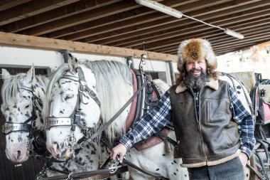 Man wearing a fur hat and leather vest stands in front of two white horses in a stable, holding the horse's reins and smiling. | © Kleinwalsertal Tourismus eGen • Photographer: Dominik Berchtold